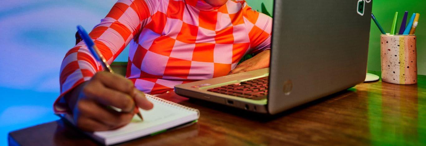 um notebook em cima de uma mesa e uma pessoa escrevendo um resumo sobre citologia em um caderno que está do lado do notebook