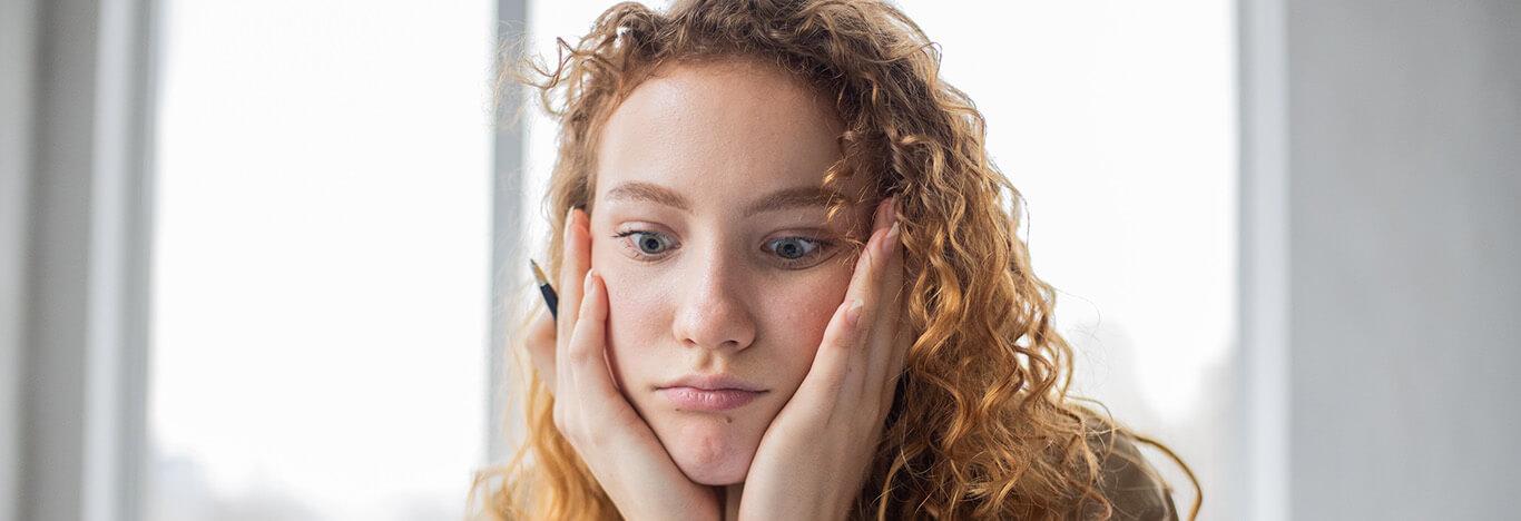 menina com cada mão de um lado da bocheca, com um lápis entre os dedos de uma mão e olhando para baixo pensando se pode ir ao banheiro durante a prova do enem